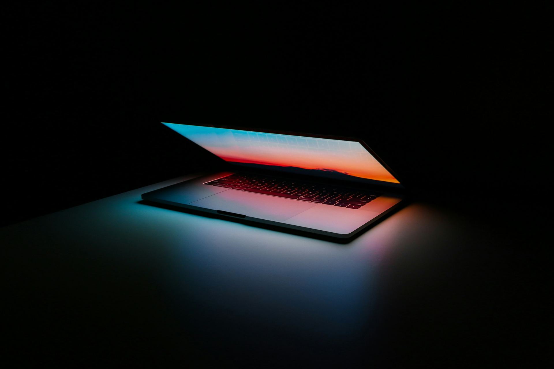 A laptop opening in a dark room, its screen casting a warm ember glow across the desk