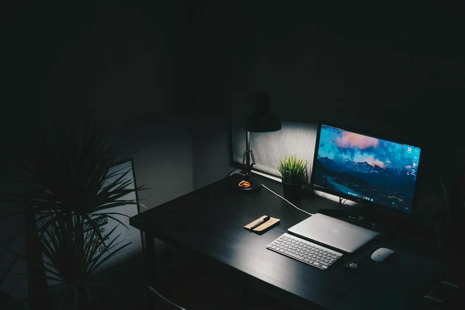 A dark workspace lit by a single lamp, a monitor and laptop on the desk