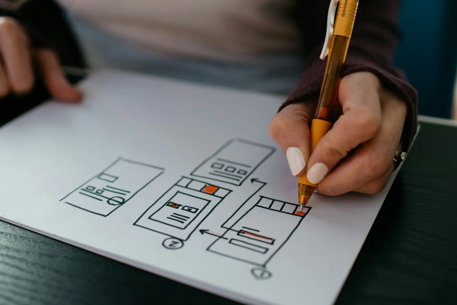 A hand sketching an interface wireframe on paper with an orange pen